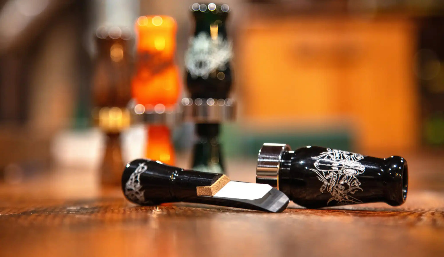 A black duck call disassembled to show its reed and barrel, with other calls blurred in the background.