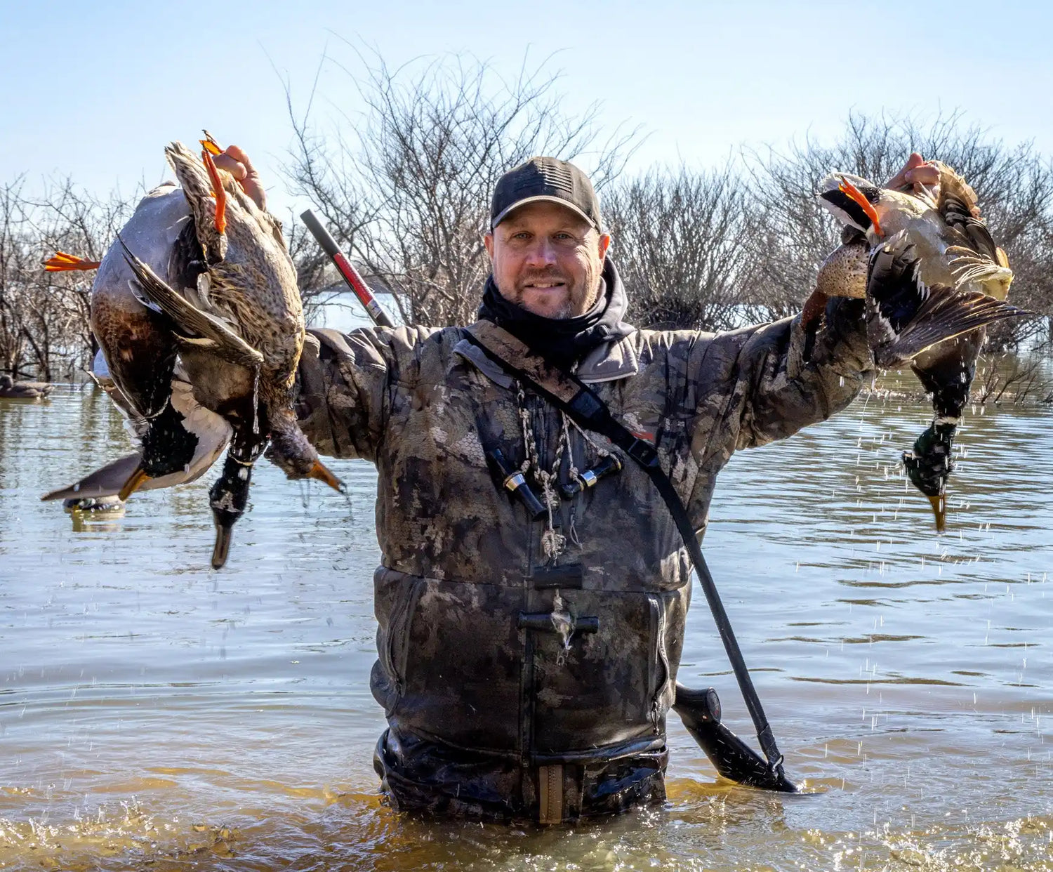 A smiling hunter in waders holding several freshly hunted ducks.
