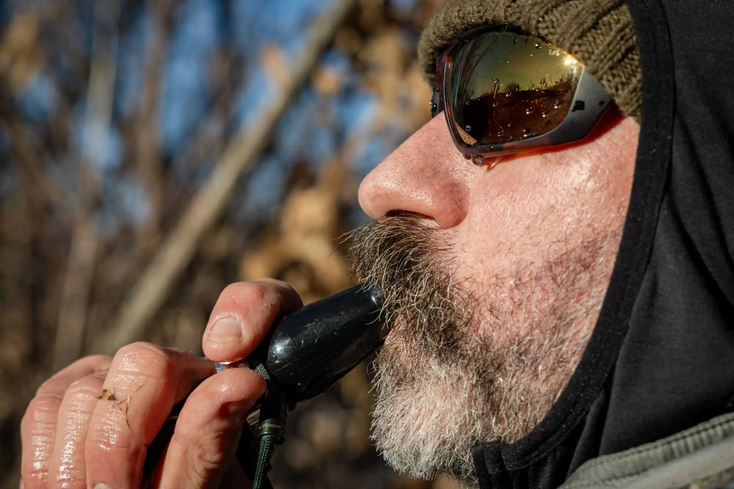 A person with a beard and sunglasses blowing into a dark duck call.