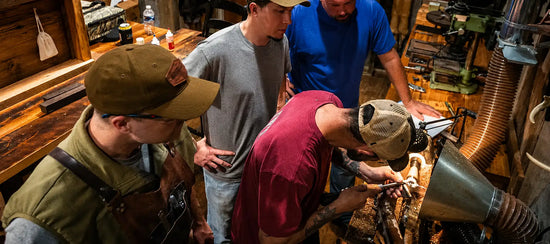A man in a red shirt and beige cap is focused on carving wood with a hand tool, surrounded by workshop tools and equipment.
