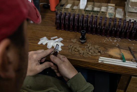 A person’s hands using a tool to cut small, white, rectangular pieces of material on a wooden surface, surrounded by other small, brown, irregularly shaped pieces and rows of dark bottles.