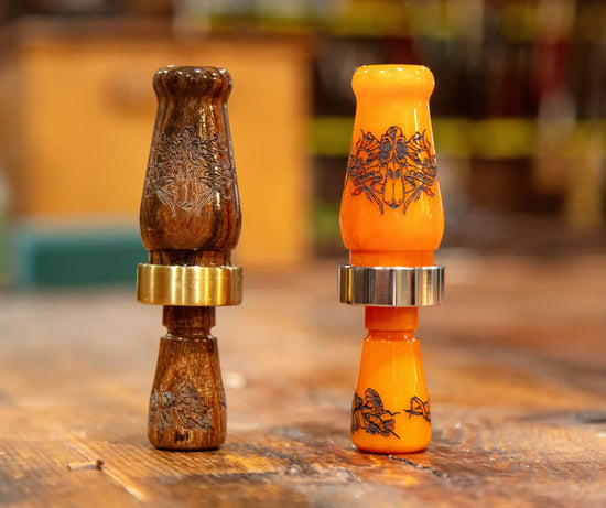 Two elaborately decorated duck calls, one in dark wood with brass accents and the other in bright orange with silver accents.